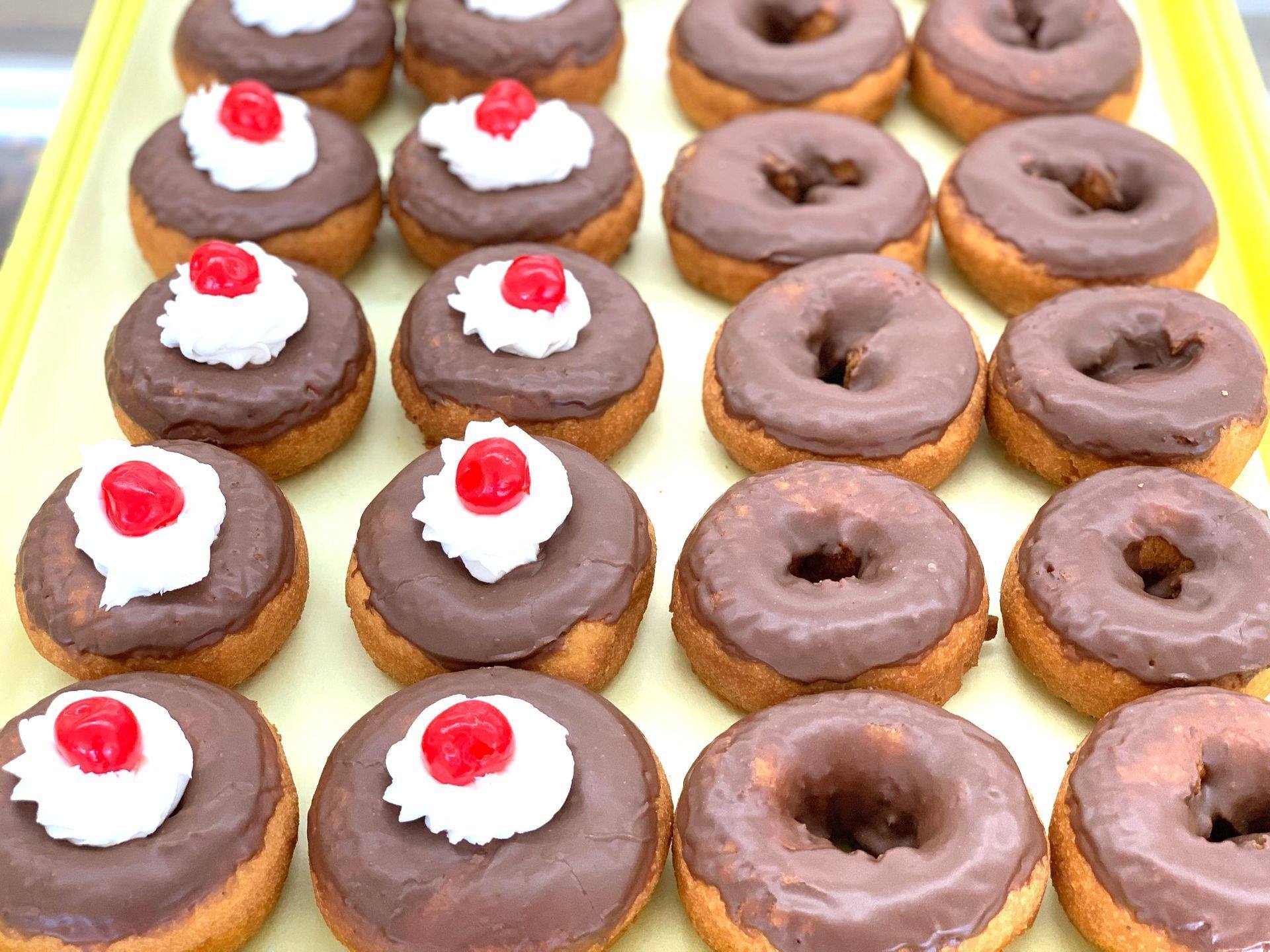 Trays of chocolate-glazed cake donuts, some topped with whipped cream and a cherry